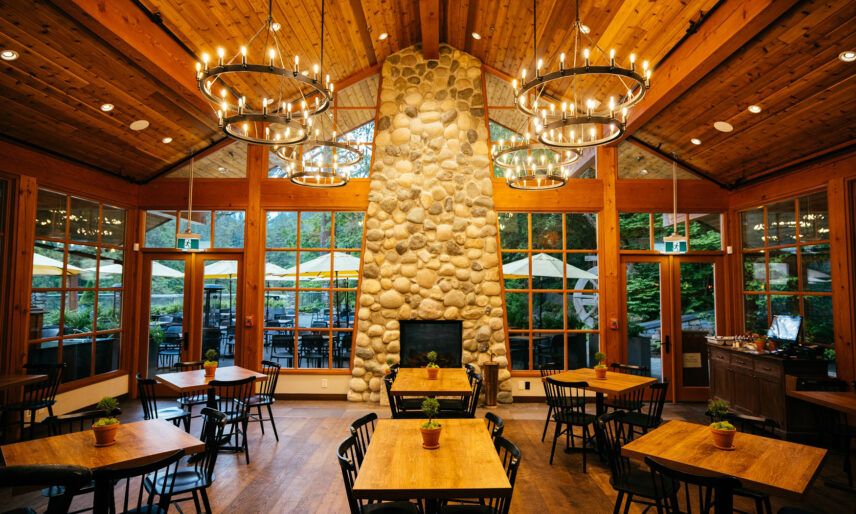 The interior of the Cliff House Restaurant at Capilano Suspension Bridge Park features rustic wooden accents, a river rock fireplace and large windows offering scenic views of the surrounding forest, and comfortable seating, creating a cozy and inviting atmosphere for diners