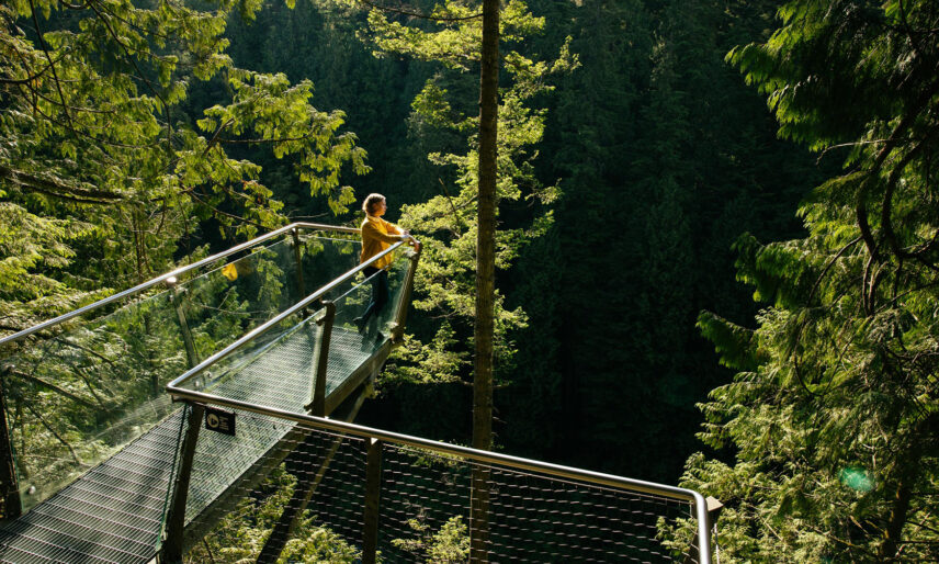 A woman enjoys the warmth of the sun while standing on Cliffwalk's long lookout platform, extending out from the cliff's edge, offering panoramic views of the surrounding natural beauty at Capilano Suspension Bridge Park