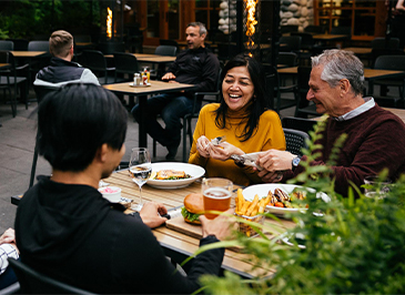 Four guests laugh and smile as they enjoy a meal on the Cliff House Restaurant's patio, surrounded by stunning views and delicious food at Capilano Suspension Bridge Park.